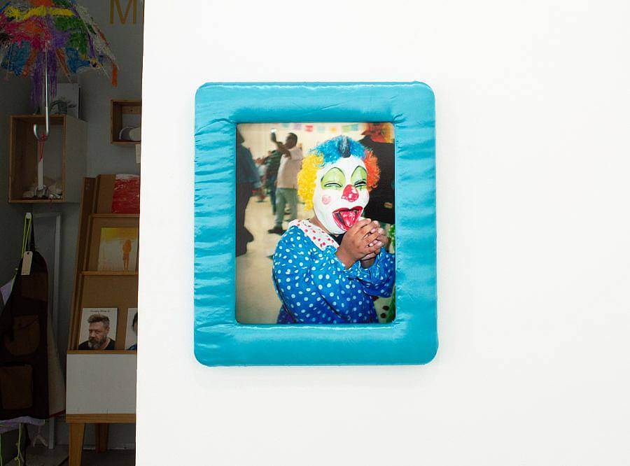 Image Description: This is a photograph of a clown in a blue satin frame. The clown has its hands grasped in from of it and is wearing a multi-colored wig and mask. The clown is wearing a blue and white polka dot outfit.