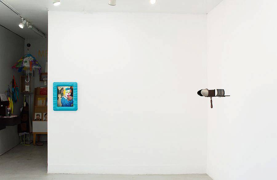 Image description: This is an installation shot of the gallery. Straight again, close to the edge of the wall is an image of a clown by Pamela Ramos. Hanging in front of the other wall to the right is a set of binoculars looking at a negative.