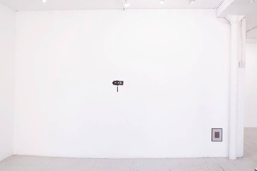 Image Description: This is an installation shot of the gallery. Hanging in front of the center of the wall is a set of hanging binoculars pointed at a negative image. Hung near the floor in the bottom left corner, is an image of the gallery. Both pieces are by Kioto Aoki.